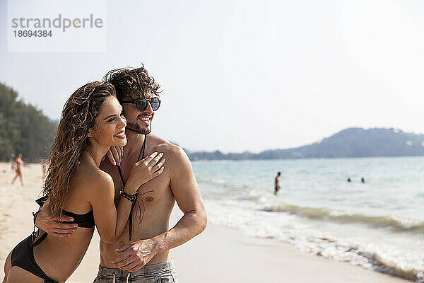 Lächelnde junge Frau umarmt ihren Freund am Strand