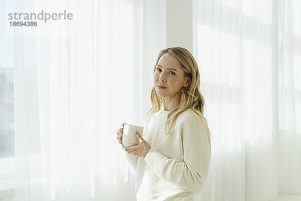 Blonde Frau hält Kaffeetasse vor durchscheinendem Vorhang