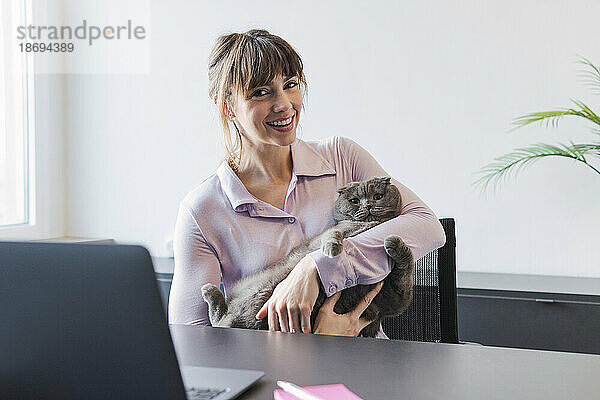Glückliche Geschäftsfrau hält Katze am Schreibtisch sitzend