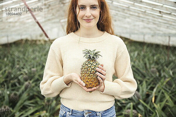 Lächelnde rothaarige Frau hält Ananas im Gewächshaus