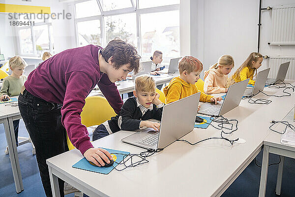 Freiwilliger hilft einem Jungen  der mit Freunden in der Schule am Schreibtisch sitzt und einen Laptop benutzt