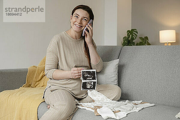Glückliche schwangere Frau  die zu Hause mit Ultraschallbildern auf dem Smartphone sitzt und spricht