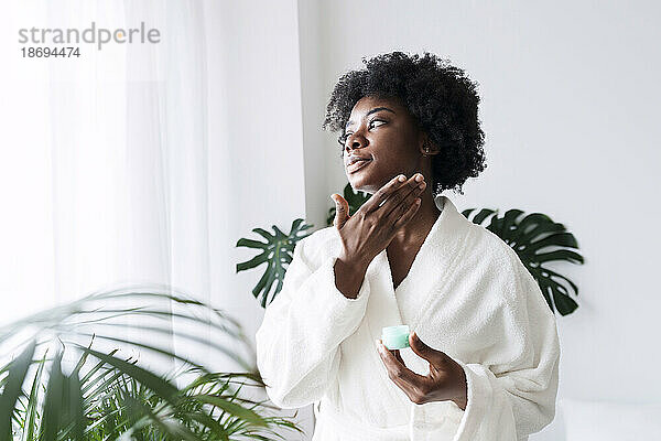 Woman wearing bathrobe applying moisturizer on face at home