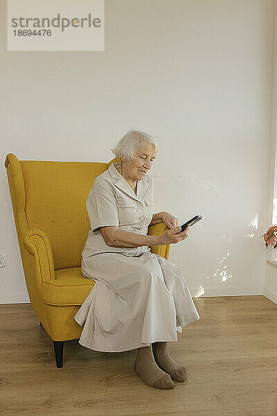 Ältere Frau sitzt zu Hause im Sessel und nutzt ihr Smartphone