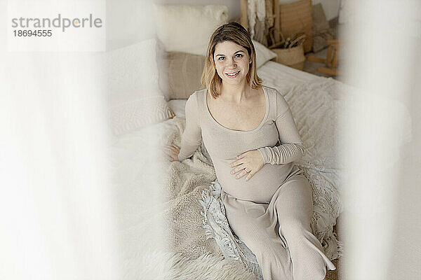 Lächelnde schwangere Frau  die zu Hause auf dem Bett sitzt und durch den Vorhang gesehen wird