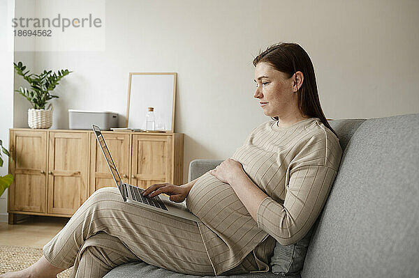 Schwangere Freiberuflerin mit der Hand auf dem Bauch arbeitet im Heimbüro am Laptop