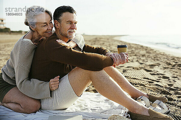 Lächelnde Frau umarmt Mann mit Einwegbecher am Strand