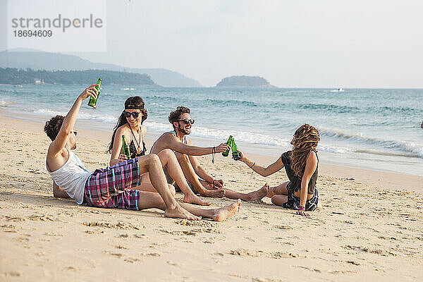 Happy friends toasting drink bottles sitting at beach