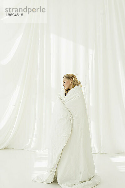 In eine weiße Decke gehüllte Frau steht neben einem durchscheinenden Vorhang