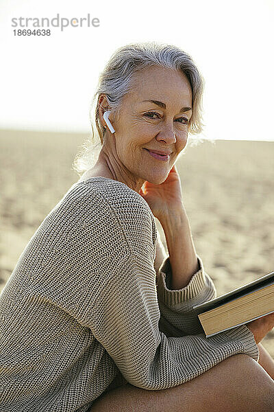 Lächelnde reife Frau mit kabellosen In-Ear-Kopfhörern am Strand