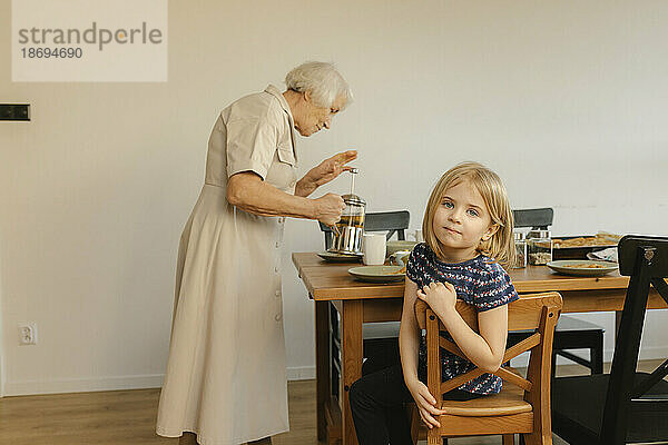 Mädchen sitzt auf einem Stuhl und Großmutter bereitet im Hintergrund zu Hause Tee zu
