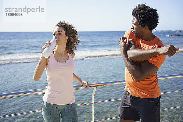 Mann trainiert mit Freundin und trinkt Wasser am Geländer in Küstengebiet