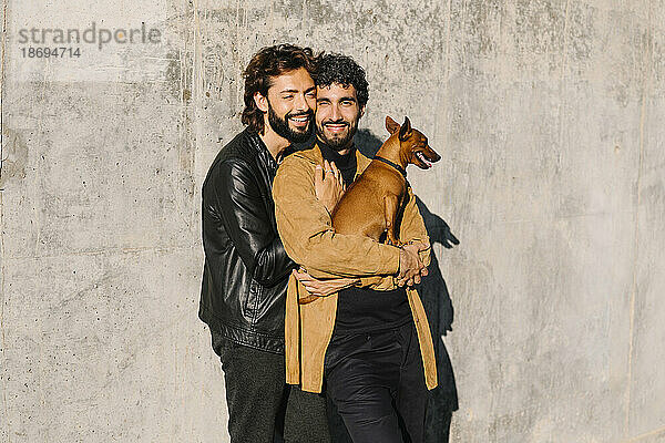 Happy gay couple with dog standing in front of concrete wall