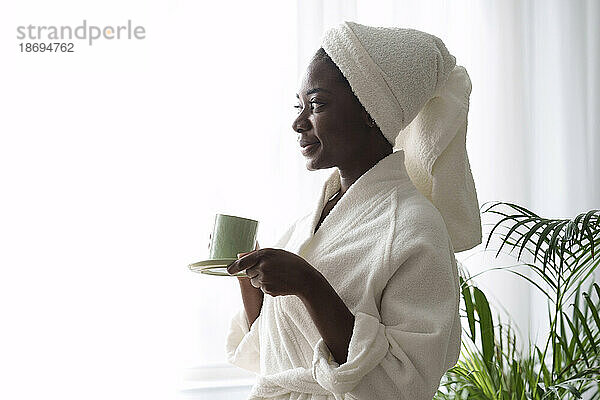 Lächelnde Frau im Bademantel hält zu Hause eine Tasse Kaffee in der Hand