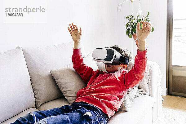 Junge mit erhobenen Armen und Virtual-Reality-Headset liegt zu Hause auf dem Sofa