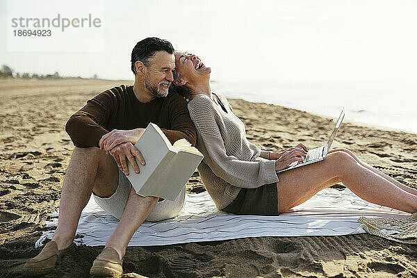 Glückliche Frau mit Laptop sitzt neben Mann und hält Buch am Strand