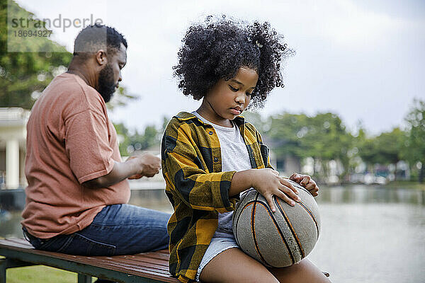 Mädchen hält Basketball und sitzt neben ihrem Vater im Park auf der Bank