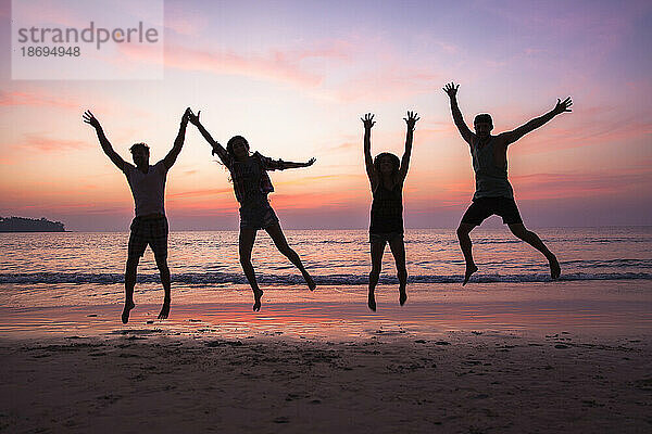 Freunde springen bei Sonnenuntergang mit erhobenen Armen am Strand