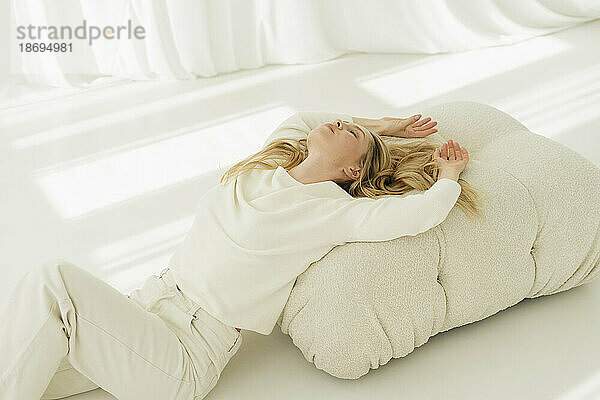 Blond woman with arms raised leaning on couch by translucent curtain