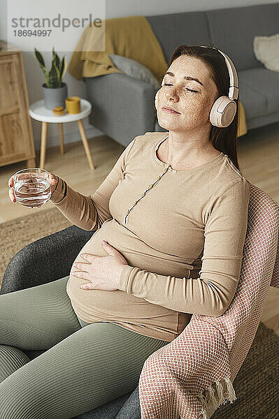 Schwangere Frau genießt Musik und hält ein Glas Wasser zu Hause