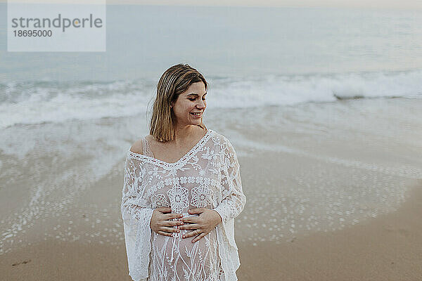 Lächelnde schwangere Frau mit den Händen auf dem Bauch steht am Strand
