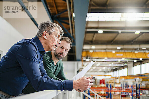 Geschäftsmann und Manager diskutieren über ein Dokument  das an einem Geländer in der Fabrik lehnt