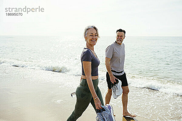 Lächelnde Frau geht mit Mann mit Sportschuhen am Strand spazieren