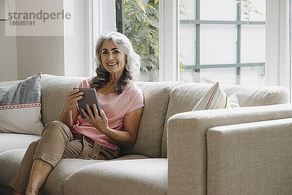 Porträt einer lächelnden reifen Frau mit Tablet  die zu Hause auf der Couch sitzt