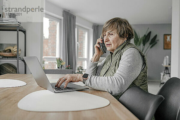 Ältere Frau benutzt Laptop und spricht zu Hause mit dem Smartphone