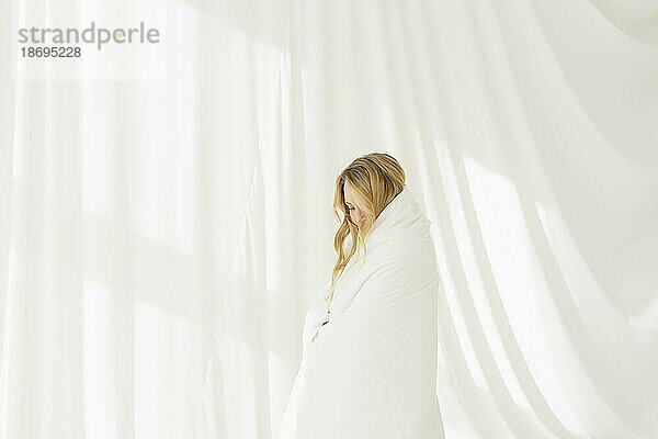 Frau  in eine weiße Decke gehüllt  steht neben einem durchscheinenden Vorhang