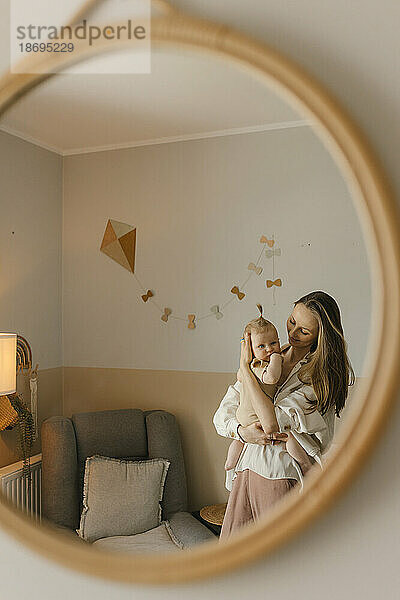 Smiling mother carrying baby daughter reflecting in mirror at home