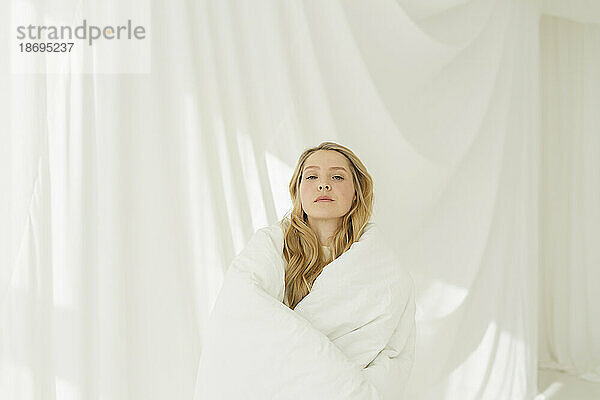 Blonde Frau  bedeckt mit einer weißen Decke  vor einem durchscheinenden Vorhang
