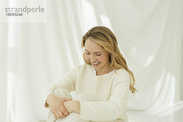 Lächelnde Frau mit blonden langen Haaren sitzt vor einem durchscheinenden Vorhang