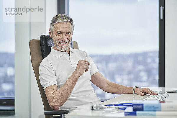Porträt eines lächelnden Arztes  der in der Arztpraxis am Schreibtisch sitzt