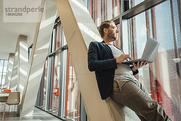 Geschäftsmann schaut aus dem Fenster und hält einen Laptop in der Hand  der sich an eine Säule im Gebäude lehnt