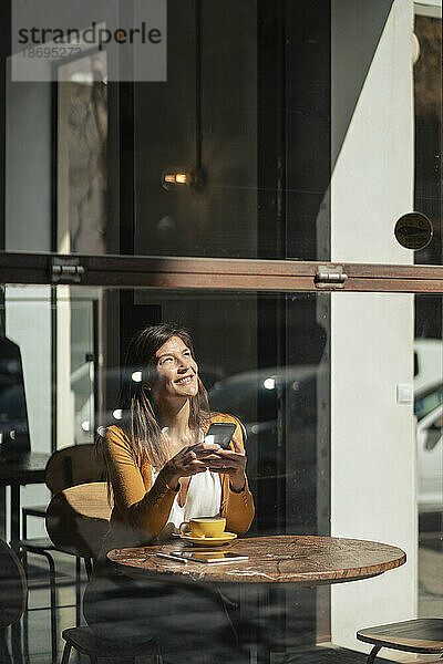 Nachdenkliche Frau mit Smartphone sitzt im Café und sieht durch Glas