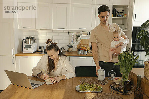 Frau arbeitet am Laptop  während Mann mit Tochter Hausarbeiten erledigt