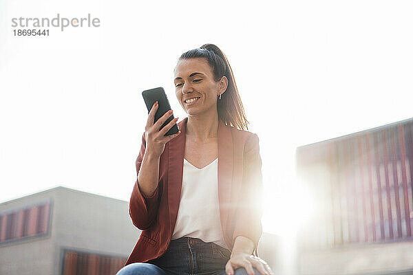 Lächelnde Geschäftsfrau  die an einem sonnigen Tag Textnachrichten über ihr Smartphone sendet