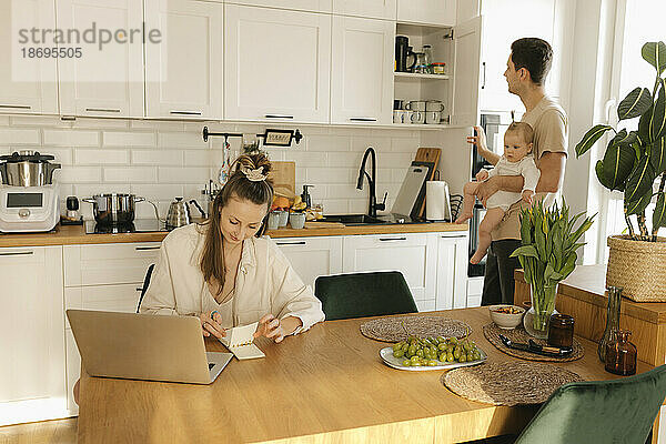 Frau arbeitet am Laptop  während Mann mit Tochter im Hintergrund Hausarbeiten erledigt