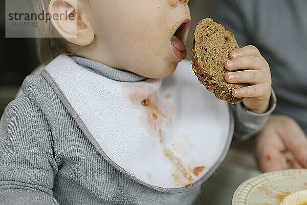 Baby boy with messy bib eating bread at home