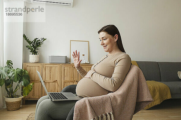 Lächelnde schwangere Frau winkt zu Hause per Videoanruf über Laptop