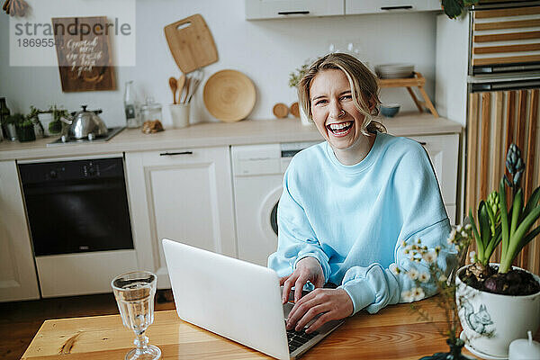 Junge Frau lacht mit Laptop am Esstisch zu Hause