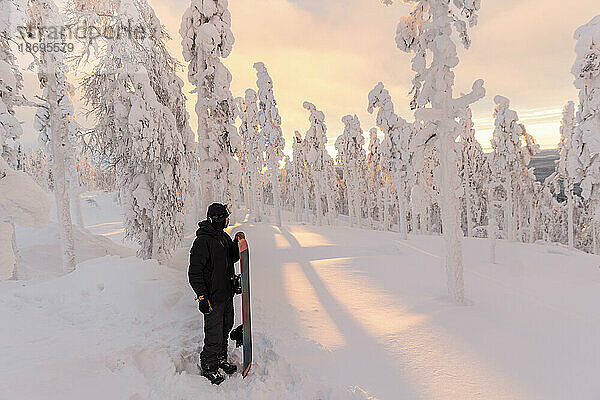 Junger Mann mit Snowboard steht in schneebedeckter Landschaft