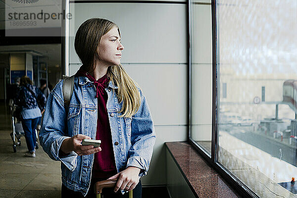 Junge Frau mit Smartphone blickt am Flughafen aus dem Fenster