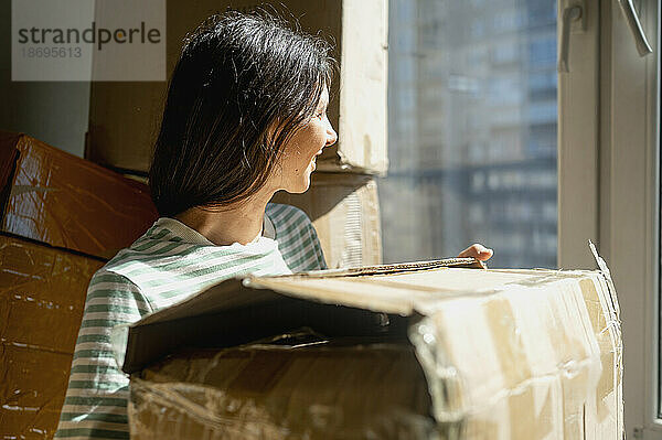 Lächelnde Frau schaut aus dem Fenster und hält zu Hause einen Karton in der Hand