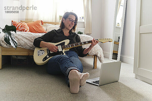 Frau lernt Gitarre und schaut sich zu Hause auf dem Laptop ein Tutorial an