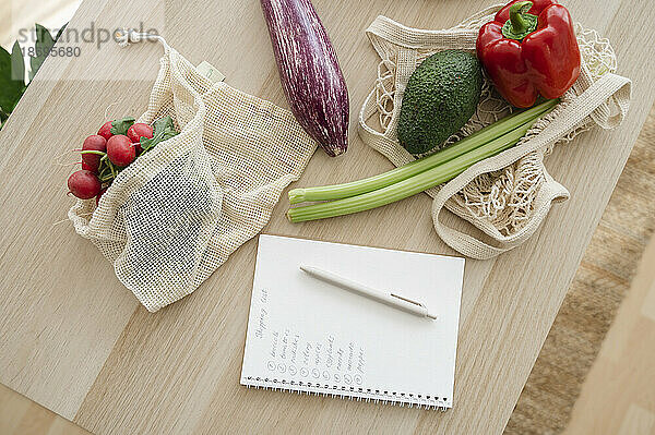 Einkaufsliste mit frischem Gemüse in wiederverwendbaren Beuteln auf dem Tisch zu Hause