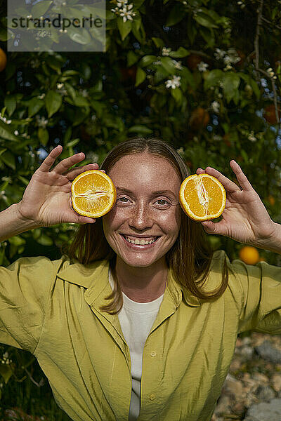 Lächelnde Frau hält Scheiben frischer Orangen im Obstgarten
