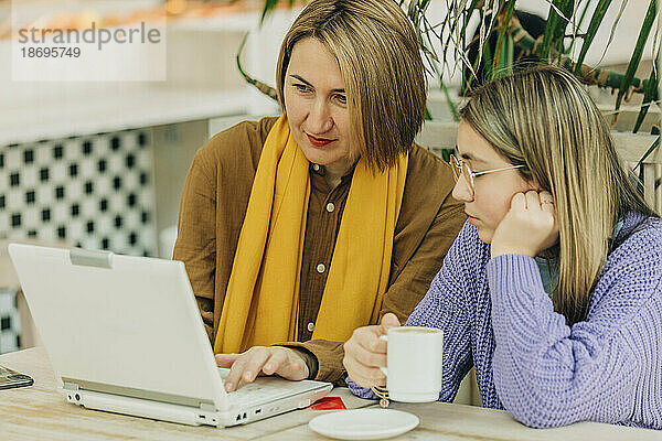 Mutter teilt Laptop mit Tochter und hält Kaffeetasse im Café