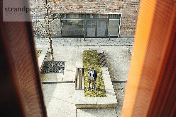 Businessman standing with laptop at office park seen through glass window
